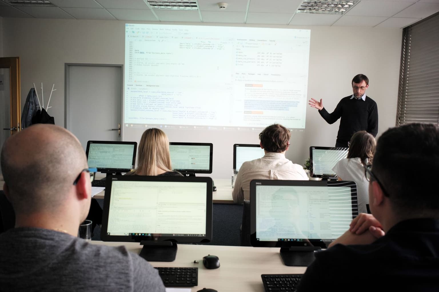 Lektor u plátna s promítaným kódem, účastníci sledují postup na svých monitorech